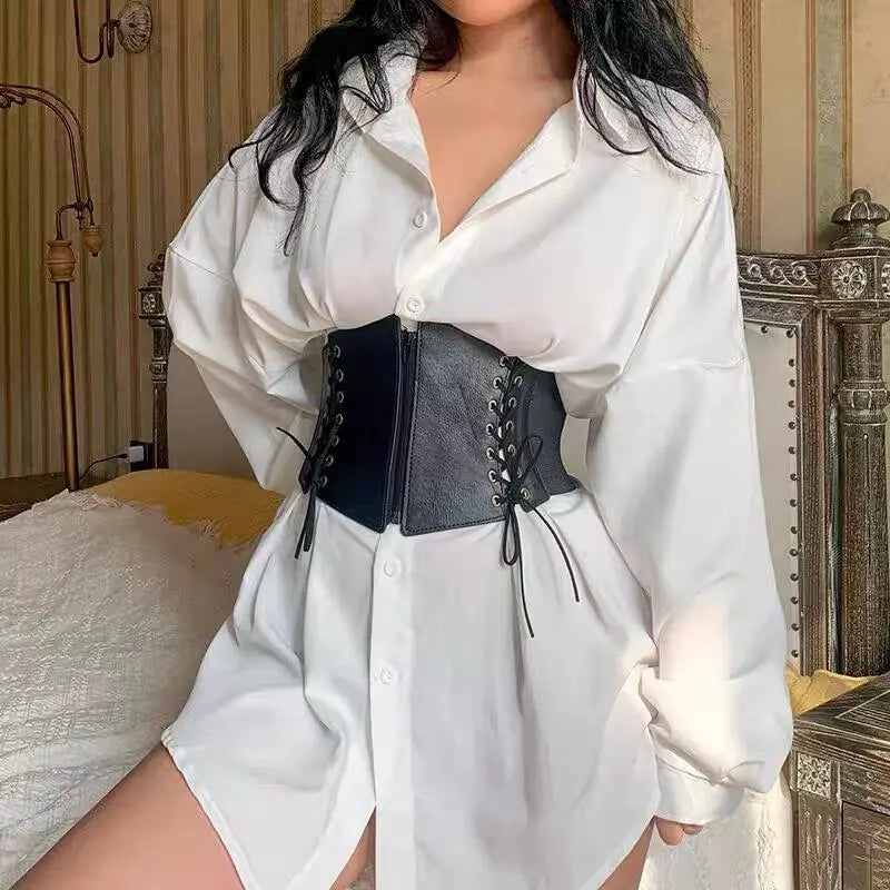 Black faux leather corset belt with antique brass grommets and lace-up sides, worn over a white button-down shirt to cinch the waist in a lifestyle setting.