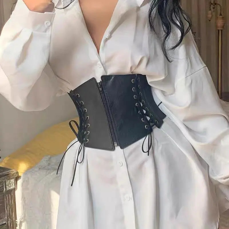 Black faux leather corset belt with antique brass grommets and lace-up sides, worn over a white button-down shirt to define the waist.