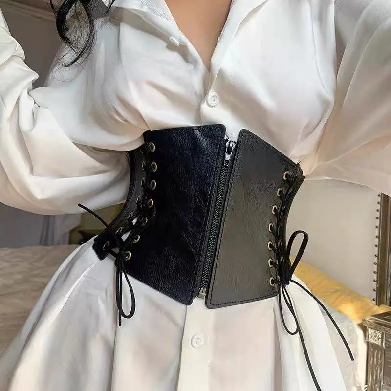 Black faux leather corset belt with antique brass grommets and lacing, worn over a white button-down shirt to define the waist.
