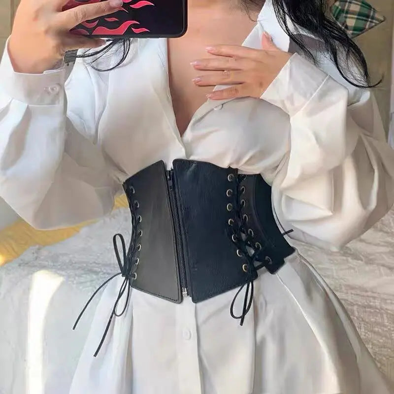 Black faux leather corset belt with antique brass grommets and lacing, worn over a white button-down shirt to cinch the waist. Product is styled in a lifestyle setting, showcasing its edgy, avant-garde appeal.