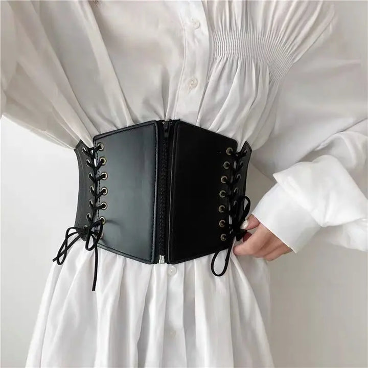 Kaelix matte black faux leather corset belt with antique brass grommets and lace-up sides, worn over a white shirt to cinch the waist.