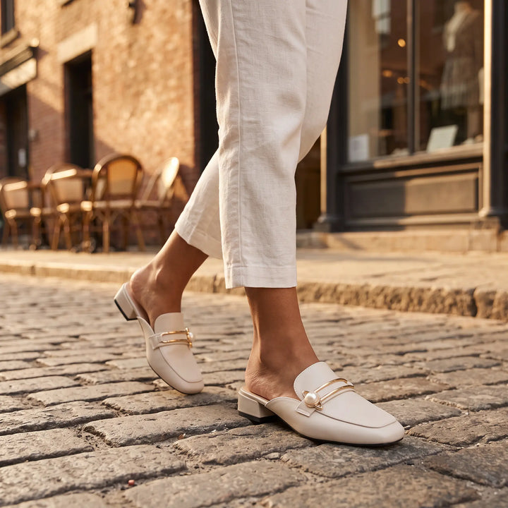 A person walks down a cobblestone street wearing light cream, square-toe mules featuring a gold bar and pearl embellishment, styled with white cropped trousers.