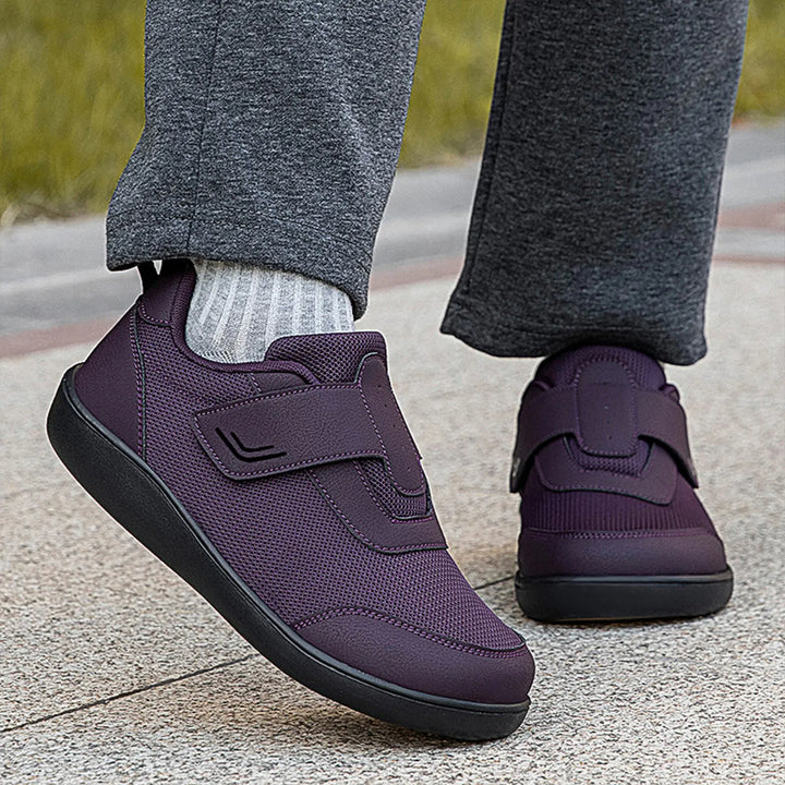 Close-up lifestyle view of purple Kaelo breathable mesh walking shoes with hook-and-loop straps, worn with grey trousers and white striped socks on a paved surface.