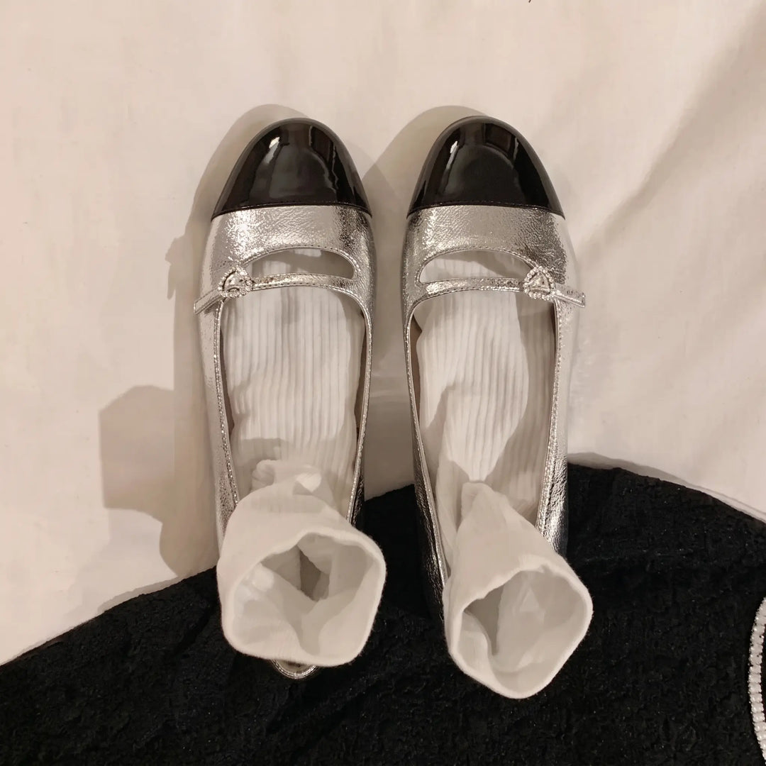 A pair of Kaelo metallic silver flat Mary Jane shoes with black patent cap toes and a delicate crystal-embellished strap buckle, presented in a studio setting with white fabric background and a hint of black garment.