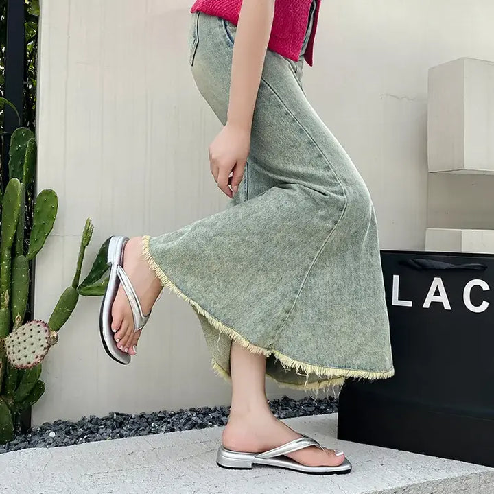 Model wearing Kaelo silver glossy PVC thong sandals with a square toe, styled with a long denim skirt in an outdoor setting.