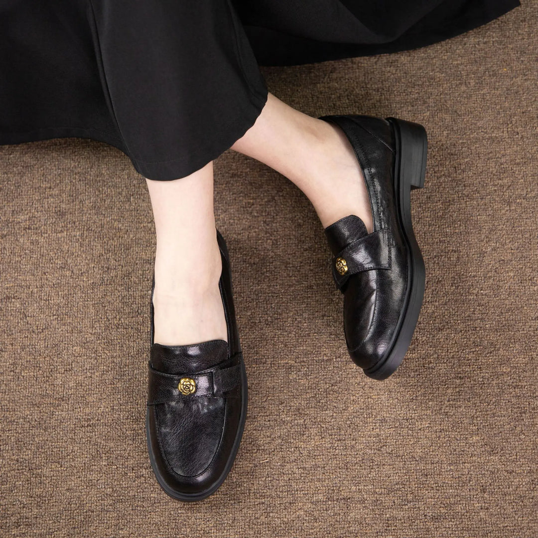 Women's black Kaelos leather loafers with a classic round toe and decorative floral buckle detail, worn by a person sitting on a textured brown carpet.