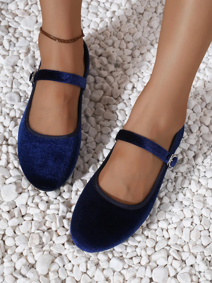 Kaelos sapphire velvet Mary Jane ballet flats featuring a round toe and an adjustable silver buckle strap, shown on feet with an anklet on a white pebble background.