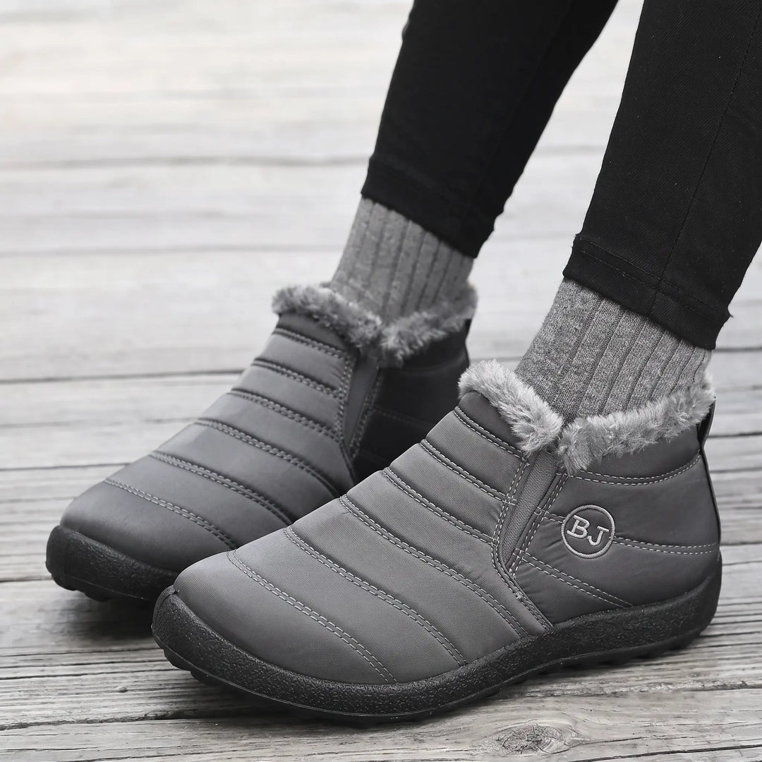 A pair of Kaelum winter warm boots in charcoal grey, featuring quilted design, faux fur trim, and elasticated sides, worn by a person on a wooden floor.