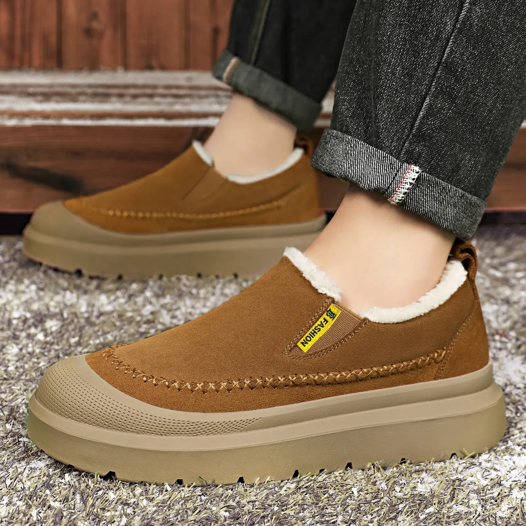 Brown men's slip-on winter boots featuring a suede-like upper, warm faux fur lining, decorative stitching, and a chunky lug sole, worn casually with cuffed jeans.