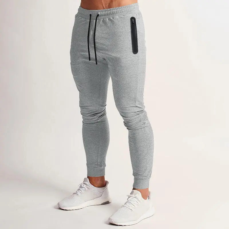 Men's heathered grey performance knit joggers featuring a tapered fit, adjustable drawstring waist, zippered side pocket, and elasticated cuffs. Model wears white sneakers.