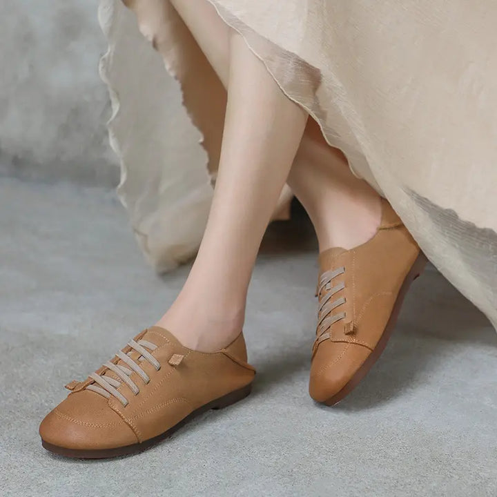 Ochre glow leather retro casual sneakers with decorative flat laces worn by a person, shown from a three-quarter front view on a grey textured studio surface.