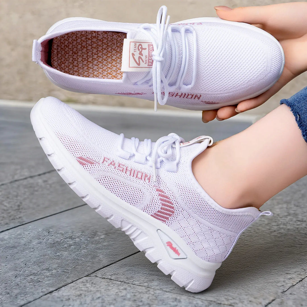 White women's breathable knit sneakers with pink "Fashion" graphic accents and lilac lining, shown worn on a foot and held by a hand against a textured grey surface.