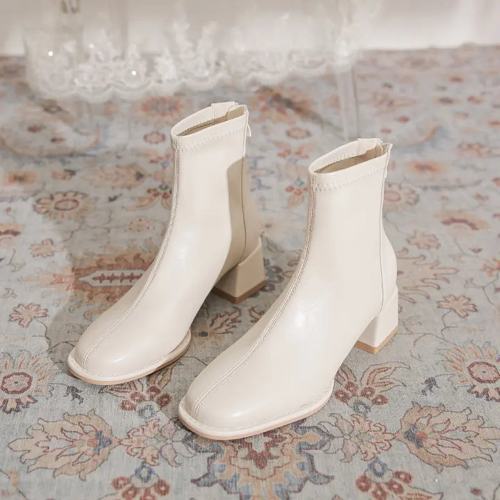 Creamy white faux leather ankle boots with a square toe, chunky block heel, and back zipper, shown from a three-quarter view on a patterned rug.