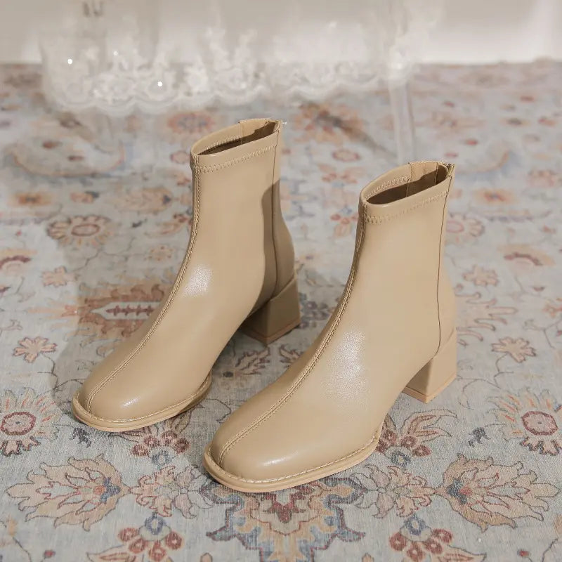 Pair of Kalyx modern chic beige ankle boots with chunky block heels and square toes, shown from the front on a decorative rug.