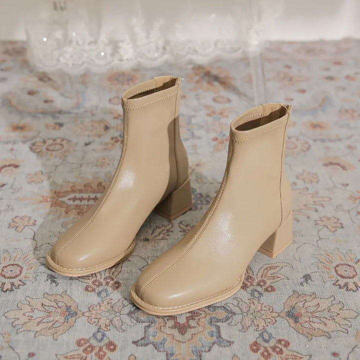 Pair of Kalyx modern chic beige ankle boots with chunky block heels and square toes, shown from the front on a decorative rug.