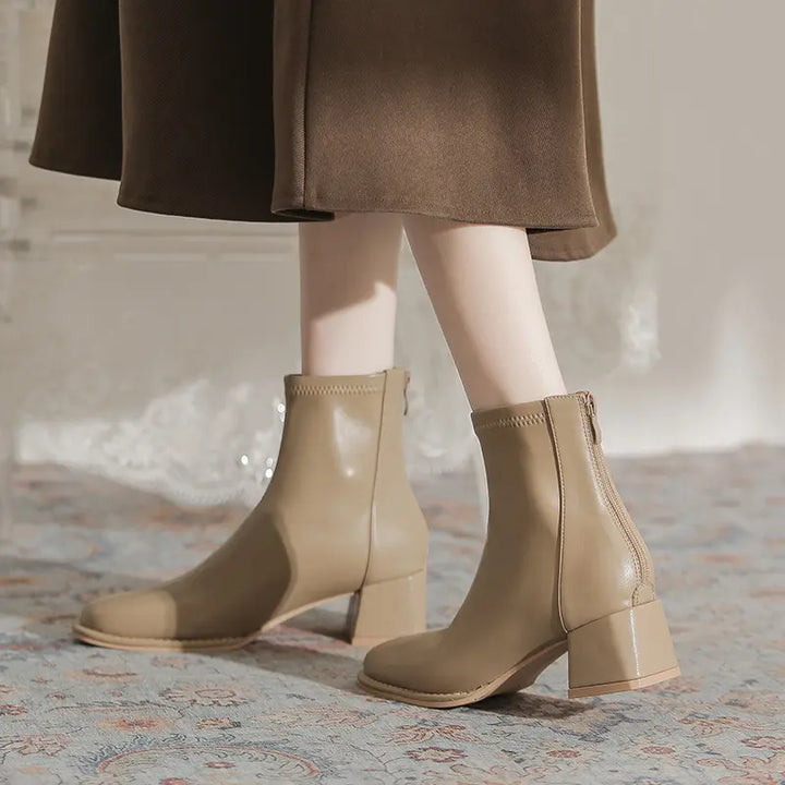 A woman wearing neutral beige Kalyx ankle boots with a chunky block heel and square toe, styled with a brown midi skirt.