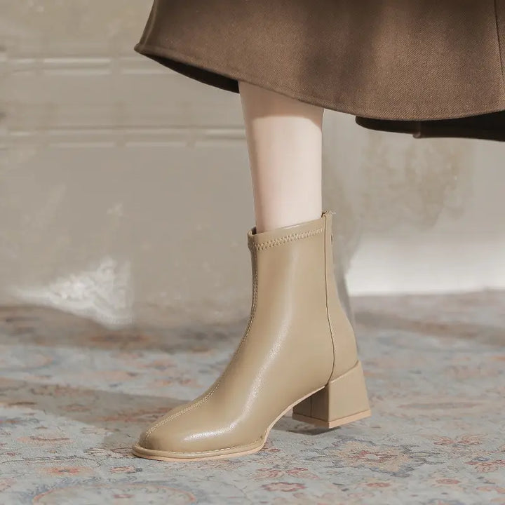 A model wears beige Kalyx ankle boots with a chunky block heel and square toe, paired with a brown skirt, showcasing modern chic style.