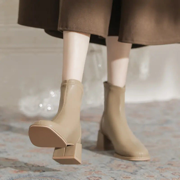 Beige faux leather ankle boots with a square toe, chunky block heel, and back zipper, shown worn by a model with a brown skirt.
