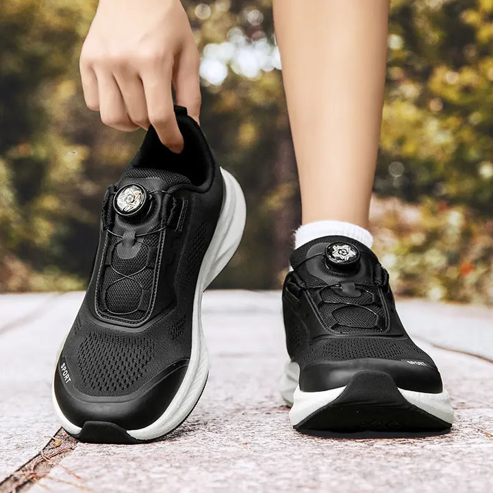 Black wide fit breathable trainers with a BOA-style closure system shown on feet with white socks, featuring a sporty design and a white EVA midsole against a blurred outdoor background.