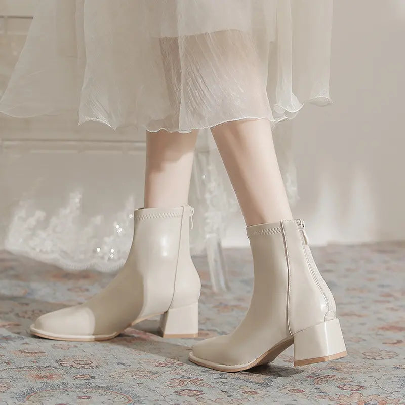 Cream faux leather ankle boots with a chunky block heel and square toe, styled with a flowing white dress and presented on a patterned rug.