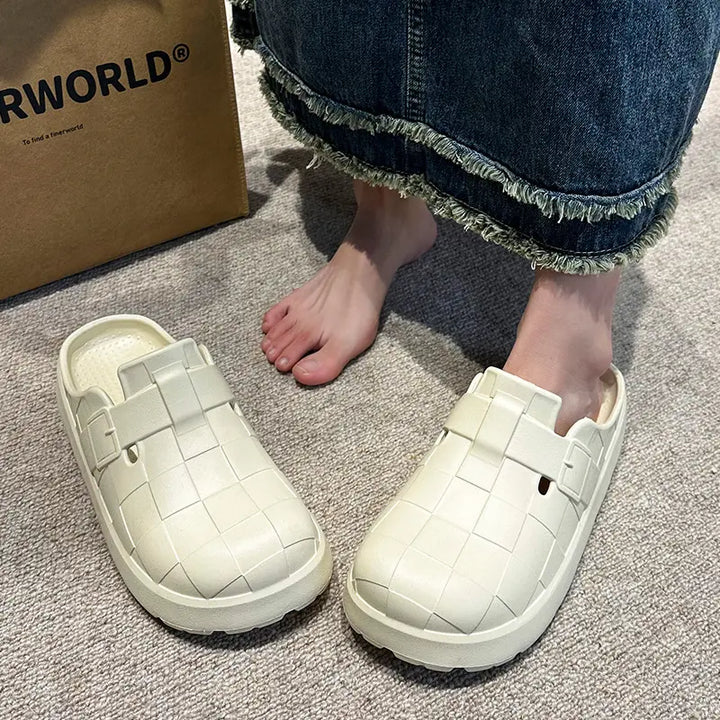 A pair of cream woven EVA clogs worn by a person in a denim skirt, shown in a lifestyle setting on a carpeted floor.