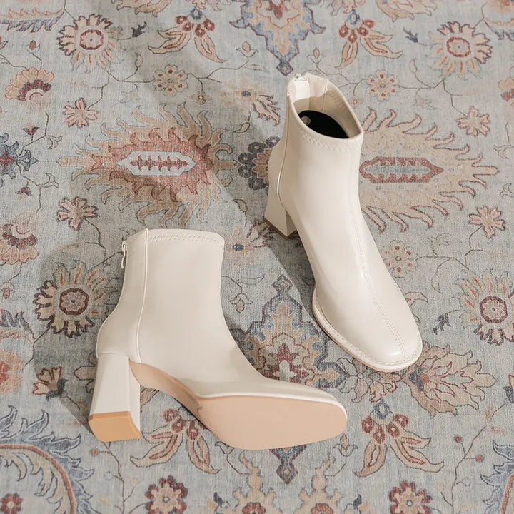Kalyx white faux leather ankle boots with a chunky block heel and square toe, shown from a three-quarter angle on a patterned rug.
