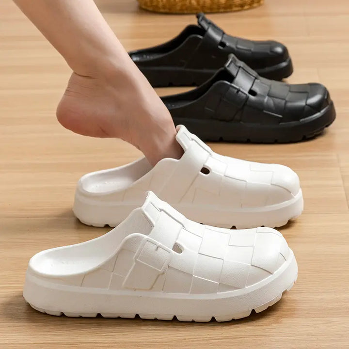A foot is inserted into a white EVA clog with a woven upper design, while black clogs are lined up behind on a wooden floor.