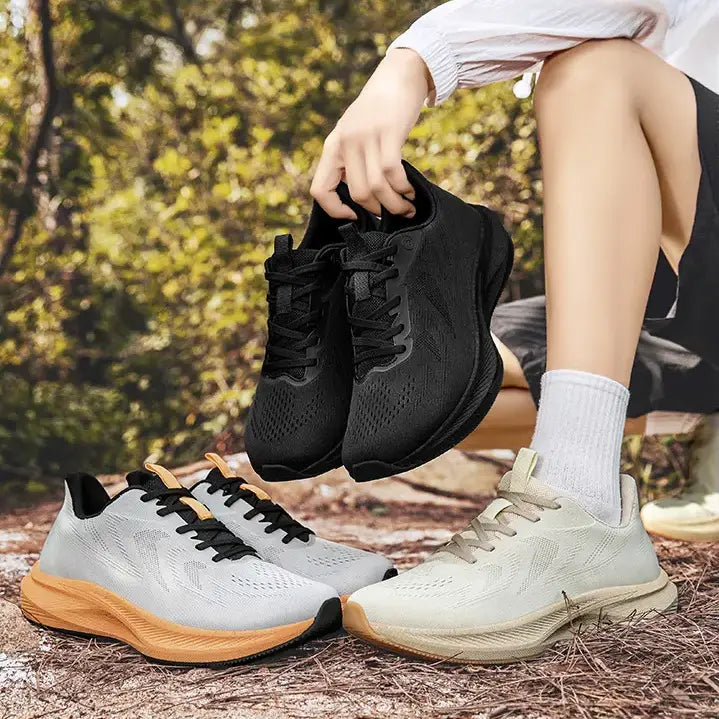 Lifestyle image of a man holding black wide-fit breathable trainers outdoors, with white and beige trainers displayed on the ground. Features a natural forest setting.