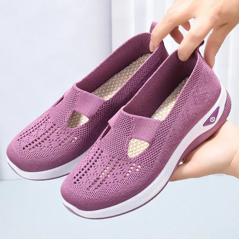 A pair of women's Kaveli amethyst breathable knit slip-on sneakers shown from a three-quarter angle, held by hands near the heel tabs, displaying their intricate weave pattern and comfortable design.