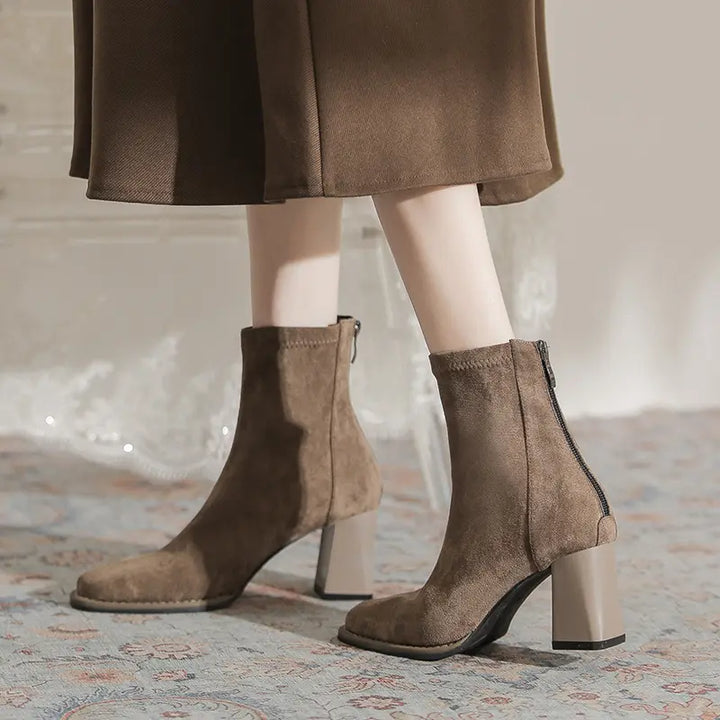 A person wearing chic khaki suede-like ankle boots with a square toe, block heel, and back zipper, styled with a brown skirt in a lifestyle setting.