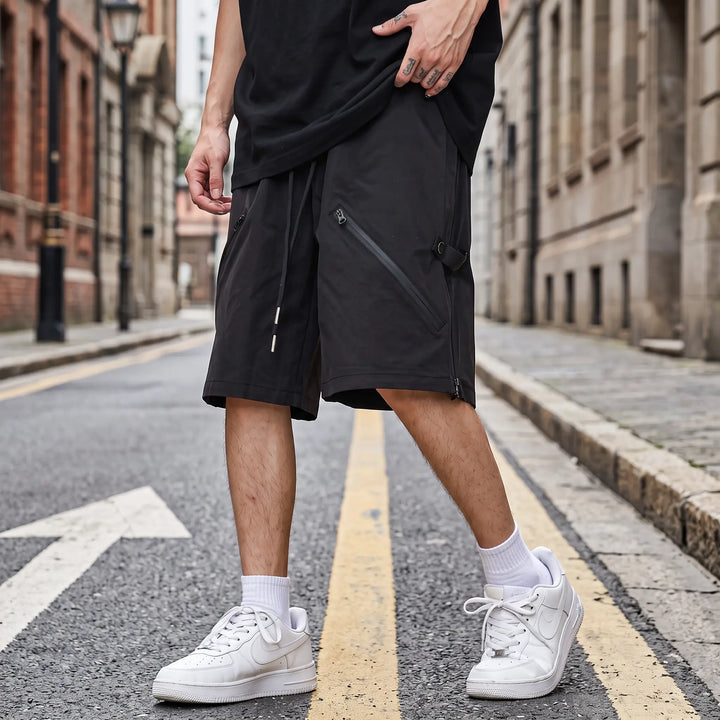 Matte black relaxed-fit technical shorts with diagonal zip pockets and flat white drawstring, shown on a model against a white studio background.