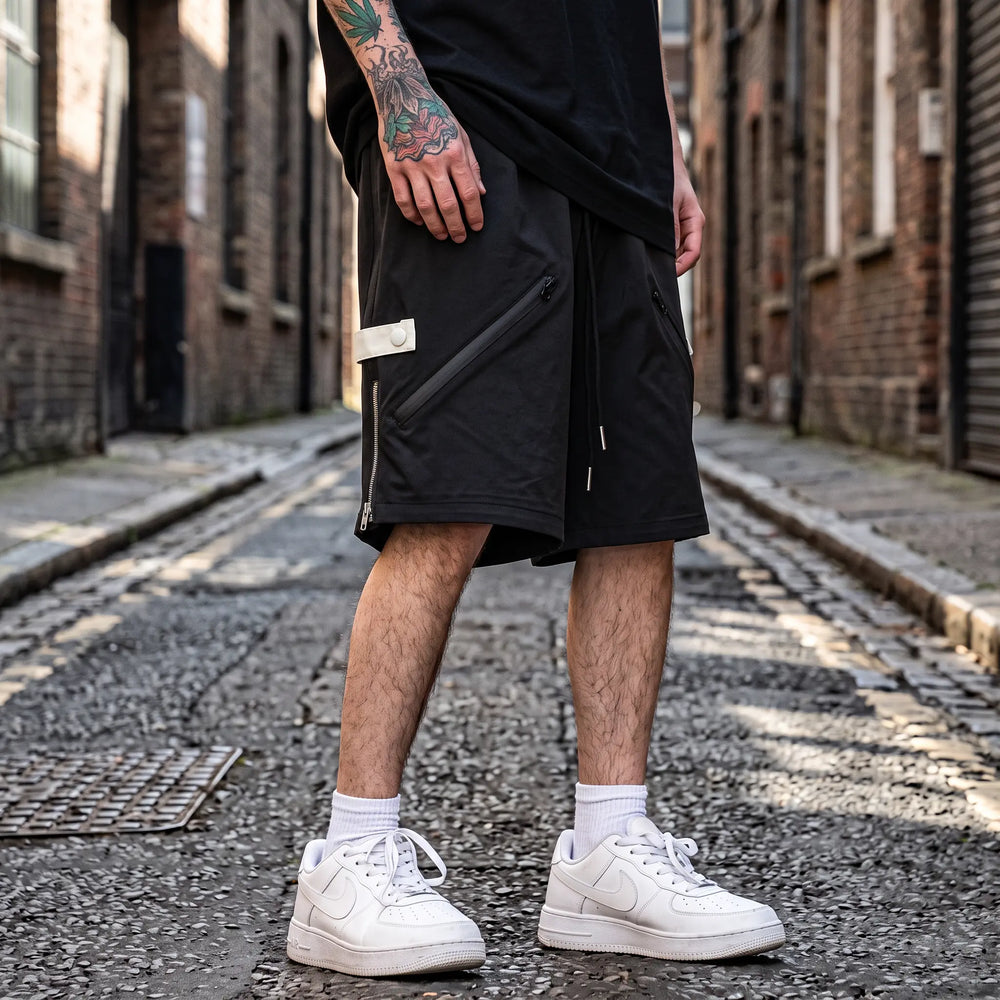 Matte black relaxed-fit technical shorts for men, featuring diagonal zip pockets and rear snap button pocket, displayed on a model against a white studio background.