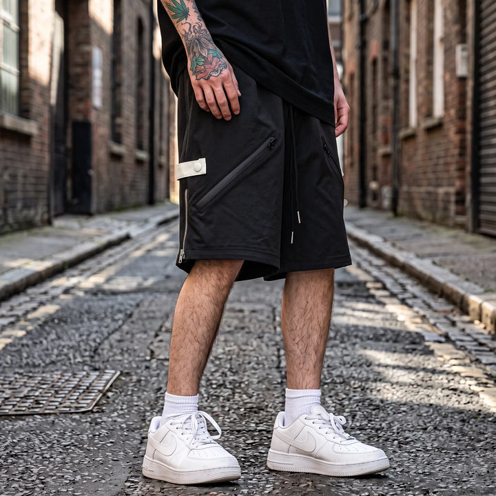 Matte black relaxed-fit technical shorts for men, featuring diagonal zip pockets and rear snap button pocket, displayed on a model against a white studio background.