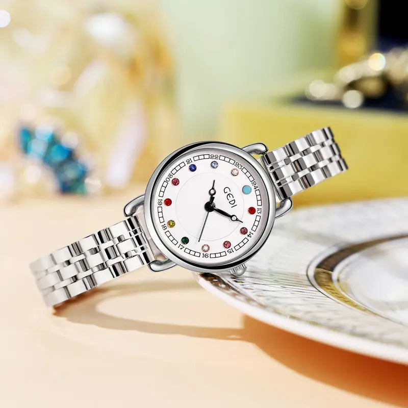 Kovian women's silver steel bracelet watch with a white dial featuring colourful gemstone hour markers, slender black hands, and a date window. Presented at a three-quarter angle on a decorative plate.
