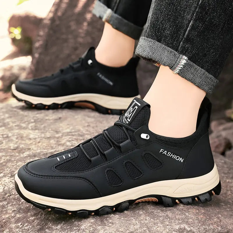 Black Krovix men's hiking sneakers with breathable mesh and synthetic upper, shown on feet in an outdoor rocky setting. Features a cushioned cream midsole and lugged outsole for all-terrain traction.