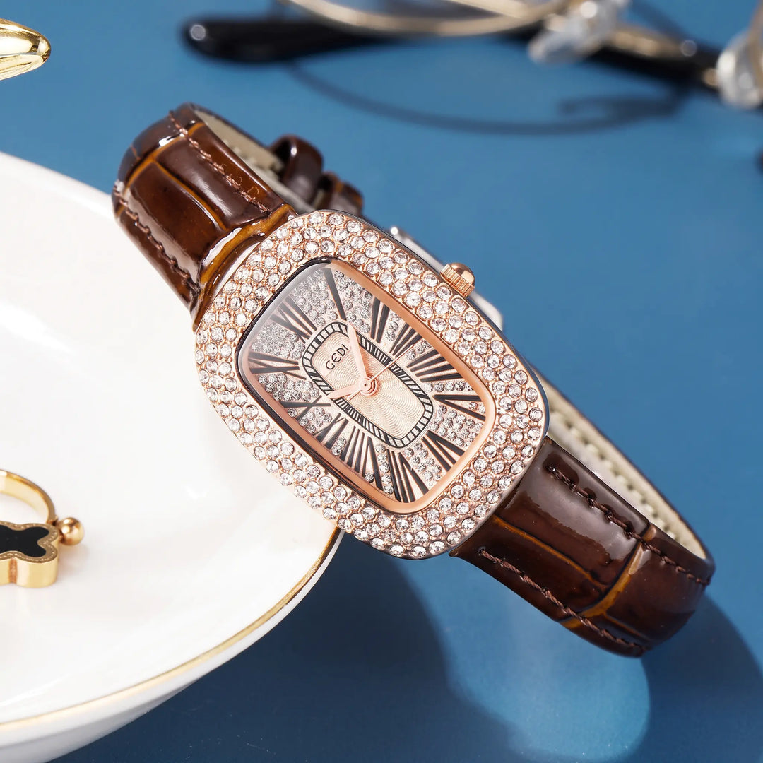 A rose gold-toned oval watch with a crystal-embellished bezel, champagne sunburst dial, Roman numerals, and a brown alligator-embossed genuine leather strap, styled on a white plate with a blue textured background.