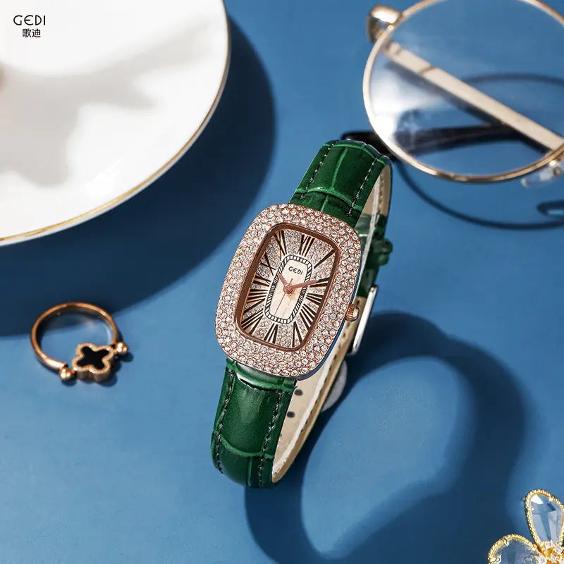 Kryllon oval luxury women's watch with a rose gold-toned crystal-embellished bezel, champagne sunburst dial, black Roman numerals, and a green alligator-embossed genuine leather strap, styled on a teal surface with accessories.