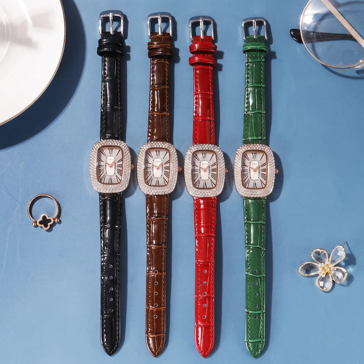 Four Kryllon oval watches with crystal-embellished bezels and alligator-embossed leather straps in black, brown, red, and green, arranged on a teal blue textured surface with styling accessories.