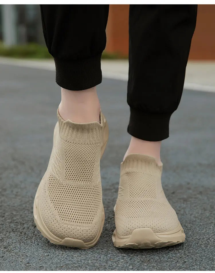 A pair of beige Kryxis knit slip-on sneakers worn by someone wearing black joggers, viewed from a front angle on an outdoor street surface, highlighting the breathable knit upper and comfortable fit.