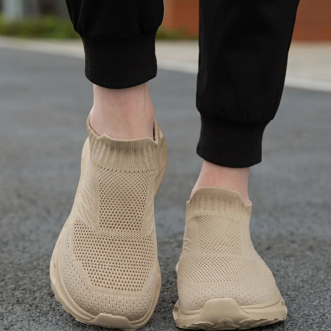 A pair of beige Kryxis knit slip-on sneakers worn by someone wearing black joggers, viewed from a front angle on an outdoor street surface, highlighting the breathable knit upper and comfortable fit.