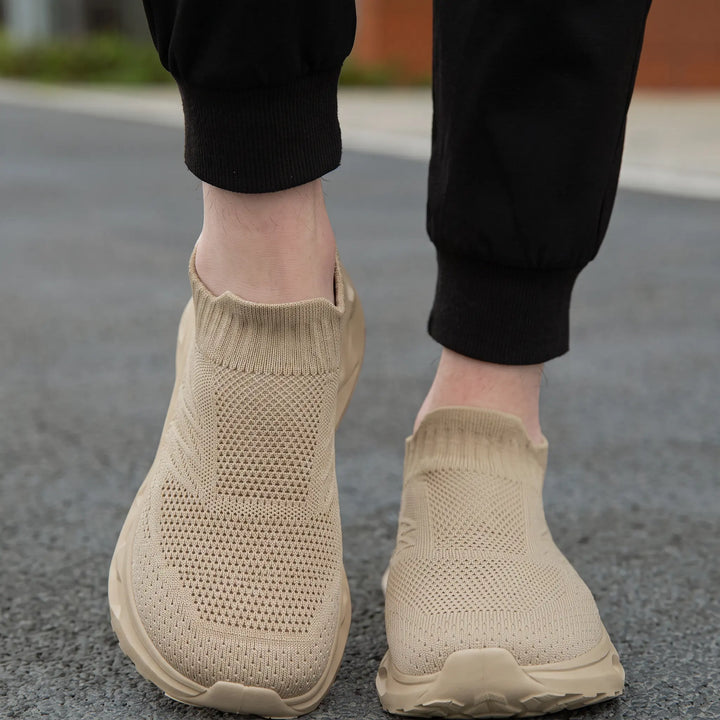 A pair of beige Kryxis knit slip-on sneakers worn by someone wearing black joggers, viewed from a front angle on an outdoor street surface, highlighting the breathable knit upper and comfortable fit.