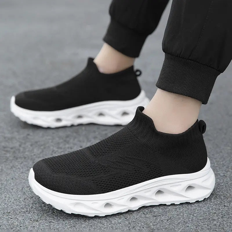 Black slip-on knit trainers with sculpted white soles, shown on feet from a three-quarter angle, emphasizing their comfortable, sock-like fit and modern athletic design.