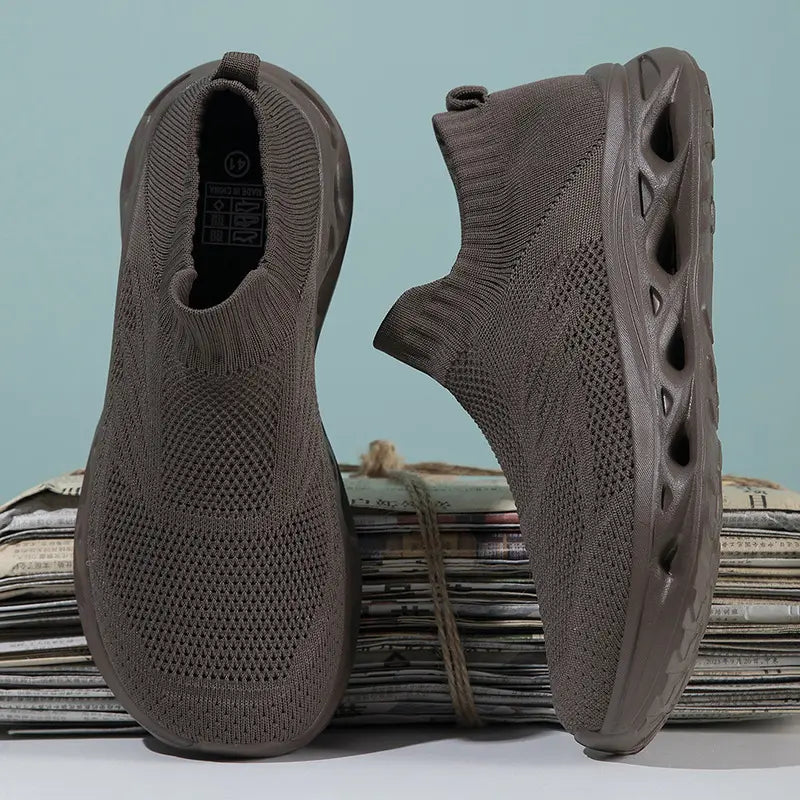 Kryxis brown knit slip-on trainers with a sock-like fit and sculpted wavy sole, shown from front and side angles on a stack of newspapers.