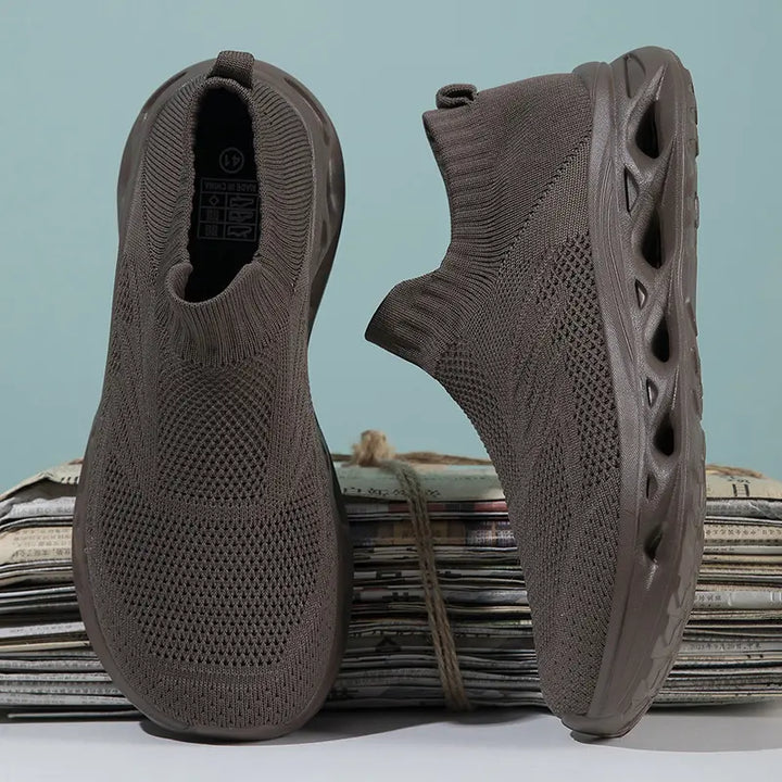 Kryxis brown knit slip-on trainers with a sock-like fit and sculpted wavy sole, shown from front and side angles on a stack of newspapers.