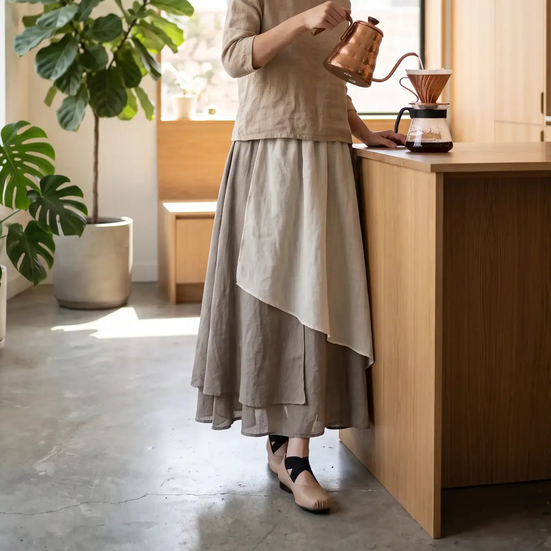 A woman wearing a flowing, layered Kumo linen midi skirt with an irregular hem in muted tonal beige, grey, and off-white, making coffee in a modern kitchen.