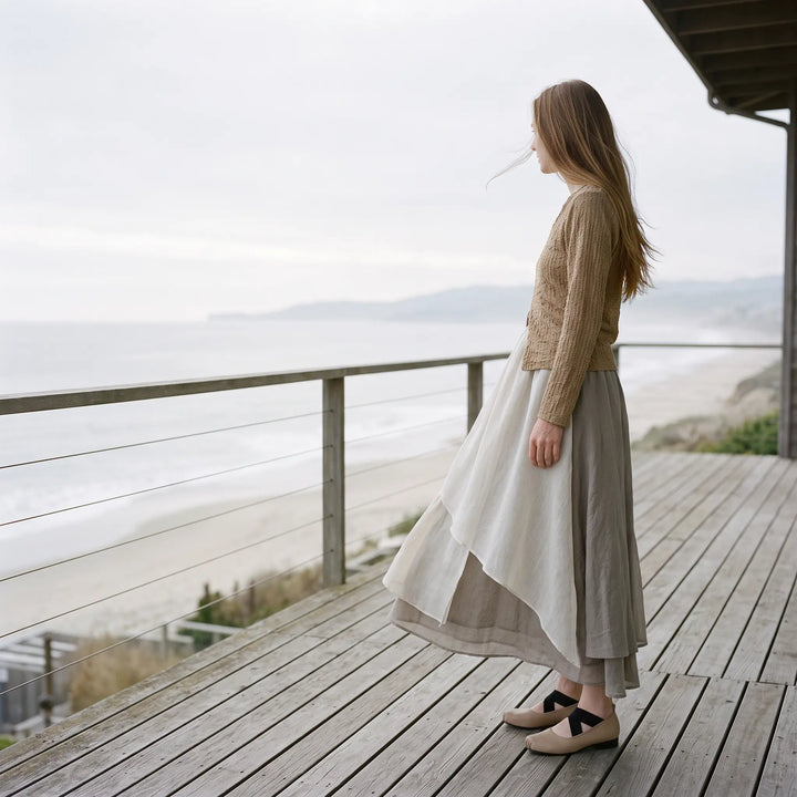 Woman standing on a wooden deck overlooking the ocean, wearing a flowing, layered midi skirt with an irregular hem and a light knit sweater.