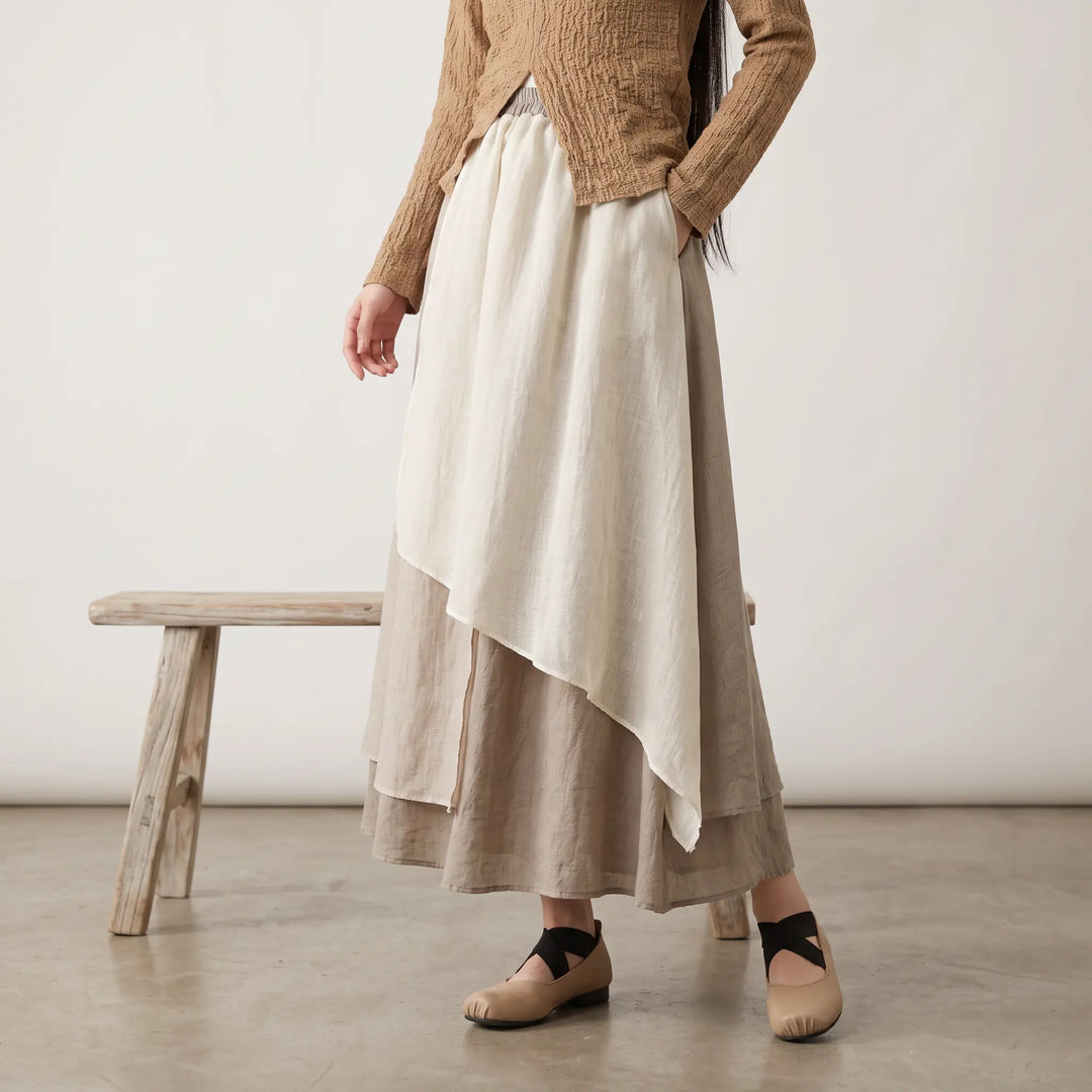 Woman wearing a flowing, ankle-length midi skirt with an irregular draped hem, featuring three distinct tonal panels of off-white, light grey-beige, and dark grey-brown linen blend fabric.