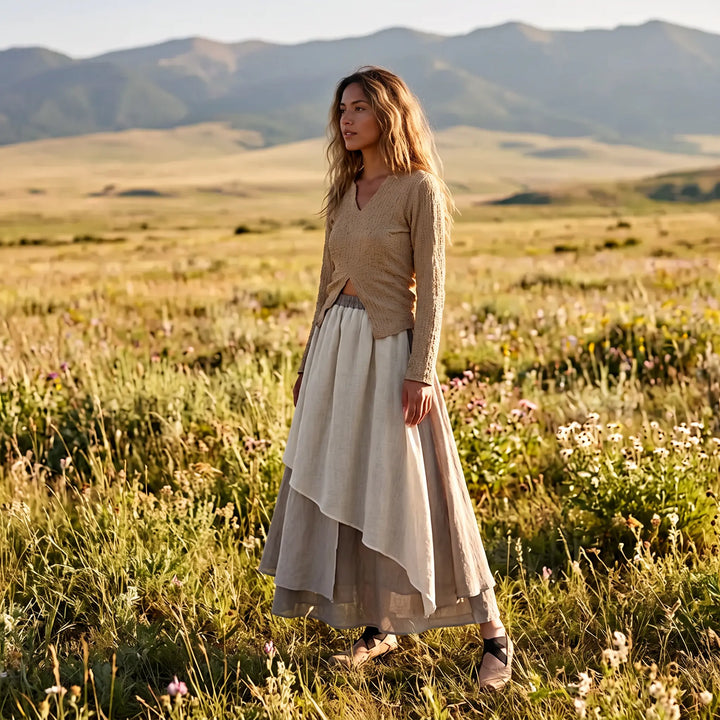 Woman in a serene meadow wearing a flowing, layered midi skirt with an irregular hem in muted tonal colors of off-white, grey-beige, and grey-brown, paired with a relaxed beige knit top.