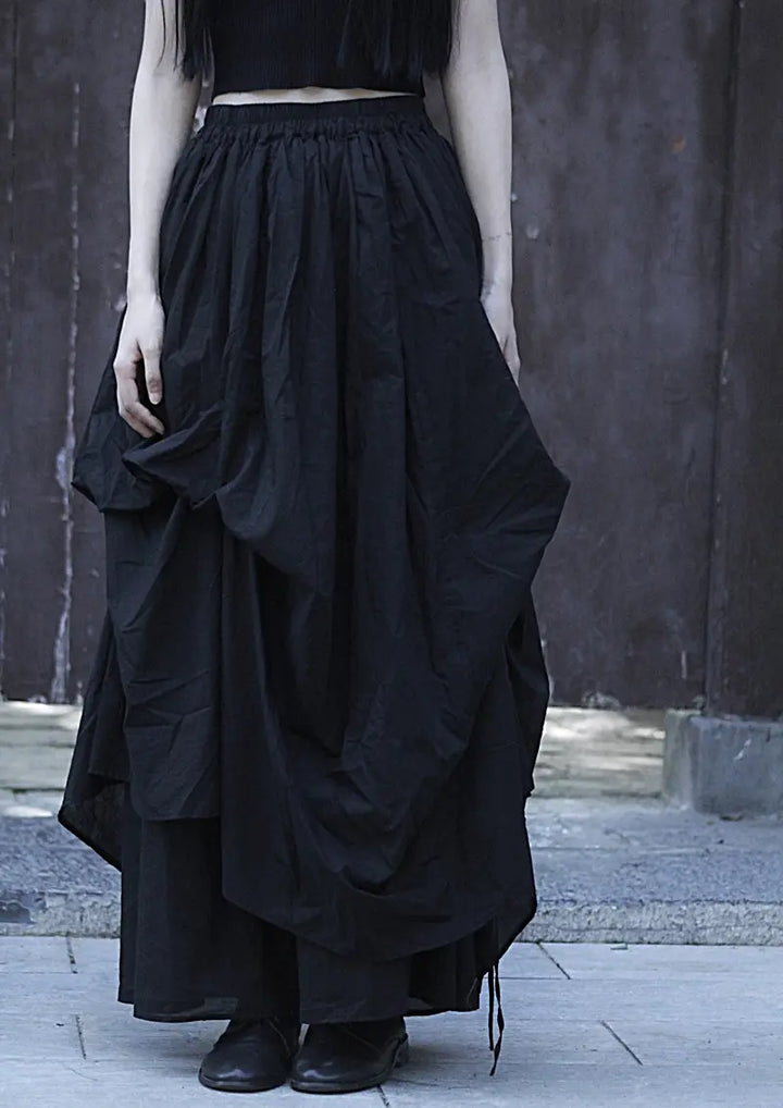 Kylori avant-garde black cotton maxi skirt with dramatic asymmetrical draping and an adjustable drawstring hem, worn by a model against a textured wall.