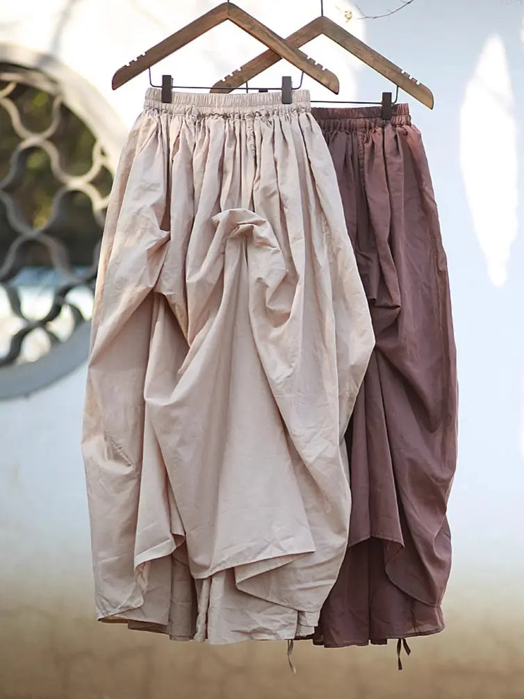 Two Kylori avant-garde maxi skirts hanging, showcasing asymmetrical draping in a light beige and a rich brown color, crafted from cotton with adjustable drawstring hems.